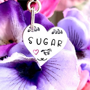May include: Silver heart-shaped charm with the word "SUGAR" engraved on it. A small pink heart and a black silhouette of a horse are also engraved on the charm.