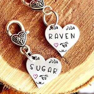 May include: Two silver heart-shaped charms with a horse and heart design. The charms are attached to silver keychains with a clip. The charms are engraved with the names "Raven" and "Sugar".