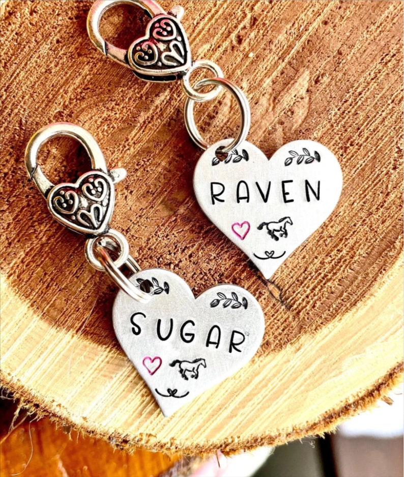 May include: Two silver heart-shaped charms with a horse and heart design. The charms are attached to silver keychains with a clip. The charms are engraved with the names "Raven" and "Sugar".