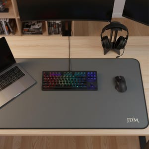 May include: A gray desk mat with a black border, a black keyboard with rainbow-colored keys, a black mouse, and a laptop. A pair of black headphones rests on a stand. The mat has the letters "JDM" in the bottom right corner.