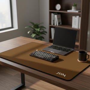 Può includere: Un tappetino da scrivania marrone con la scritta "JDM" nell'angolo in basso a destra. Una tastiera nera, un laptop e una tazza sono sul tappetino. La scrivania è in legno e si trova in un ambiente ufficio.