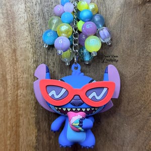 Puede incluir: Un llavero de Stitch azul con gafas de sol rojas y una bebida, sujeto a un cierre plateado y un grupo de cuentas de colores. El llavero est&aacute; sobre un fondo de madera.