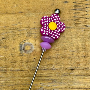 May include: A purple and white checkered flower with a yellow center on a silver metal skewer. The skewer has three purple beads and a clear plastic tip.