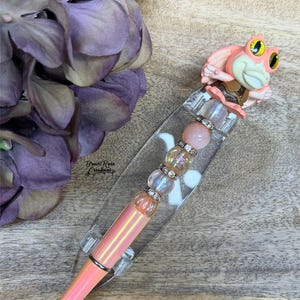 May include: A pink pen with a black tip, featuring a decorative frog figurine at the top. The pen's barrel has a pearlescent finish and is accented with clear and pink beads. The pen is displayed on a clear stand.