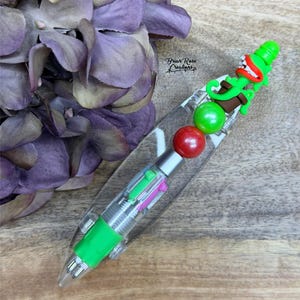 Puede incluir: Un bolígrafo multicolor con un cuerpo transparente y un agarre verde. El bolígrafo está decorado con un dinosaurio verde, cuentas rojas y verdes. El bolígrafo está sobre una superficie de madera con flores moradas en el fondo. El texto "Briar Rose Creations" es visible.
