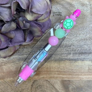 May include: A clear pen with a pink grip and a decorative turtle charm. The pen has a clear barrel with visible ink cartridges in blue and purple. The turtle charm is pink with a green shell and a pink hat. The pen is embellished with pink and green beads.