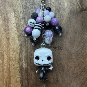 Puede incluir: Un llavero con una figura de Jack Skellington y un grupo de cuentas en negro, blanco y morado. Las cuentas incluyen diseños a rayas, con purpurina y de calaveras. El llavero tiene un cierre plateado.