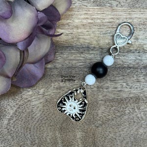 Peut inclure: Un porte-clés argenté avec un fermoir en forme de cœur et un charm en forme de soleil souriant. Le charm est noir et blanc, avec un accent de perles noires et blanches. Le texte "Brian Rose Creations" est visible.