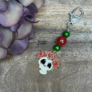 Könnte beinhalten: Ein Schlüsselanhänger zum Thema Tag der Toten mit einem weißen Totenkopf mit einem Kranz aus roten Rosen. Der Schlüsselanhänger hat grüne und rote Perlen und einen herzförmigen silberfarbenen Verschluss. Der Hintergrund ist eine Holzoberfläche.