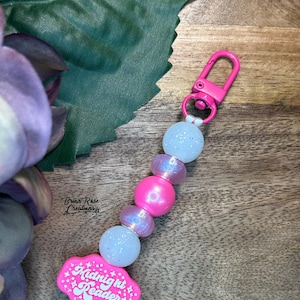 May include: A pink beaded keychain with a lobster clasp. The keychain features a cloud-shaped charm with the text "Midnight Reader", pink and iridescent beads, and a pink tassel.