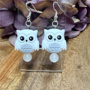 May include: A pair of dangle earrings featuring white owl charms with black eyes and light gray belly details. Each owl is suspended from a silver-toned metal hook and accented with a small, round, translucent white bead with subtle glitter.