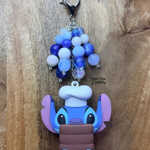 Puede incluir: Un llavero de Stitch azul y morado con un gorro de chef, sosteniendo un objeto marr&oacute;n. El llavero tiene un cierre plateado y est&aacute; adornado con cuentas azules y blancas. El texto "Brian Rose Creations" es visible.