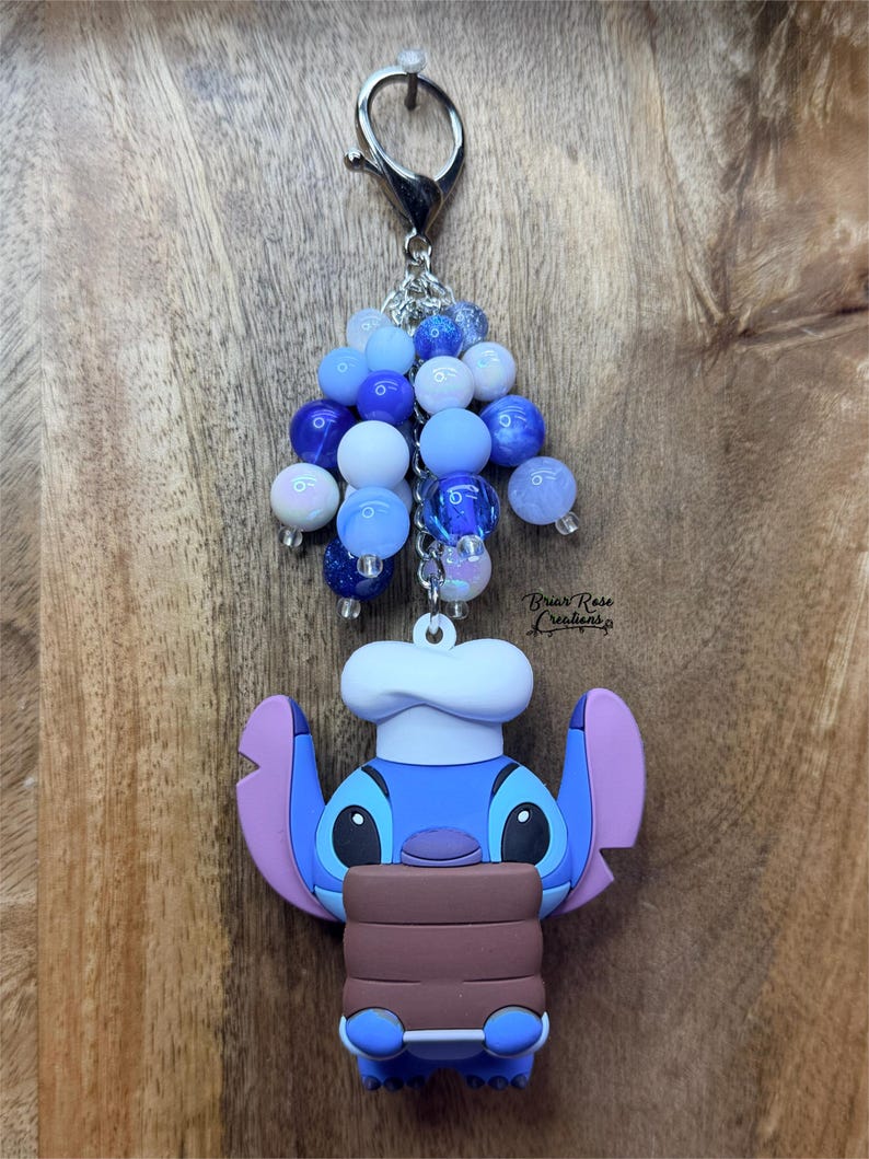 Puede incluir: Un llavero de Stitch azul y morado con un gorro de chef, sosteniendo un objeto marr&oacute;n. El llavero tiene un cierre plateado y est&aacute; adornado con cuentas azules y blancas. El texto "Brian Rose Creations" es visible.