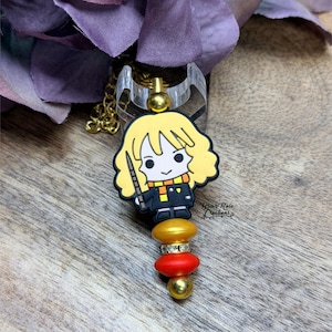 May include: A badge reel featuring a cartoon character with blonde hair, a black uniform, and a scarf. The reel has a gold chain and a clear plastic ring. The charm is attached to a series of gold, red, and gold beads.