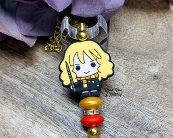 Op de afbeelding: Een badge reel met een stripfiguur met blond haar, een zwart uniform en een sjaal. De reel heeft een gouden ketting en een doorzichtige plastic ring. De bedel is bevestigd aan gouden, rode en gouden kralen.