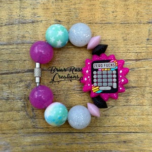 May include: A pink and black calculator charm with the text "ZERO FU*KS" on the display. The charm is attached to a bracelet made of colourful beads with glitter and star patterns.