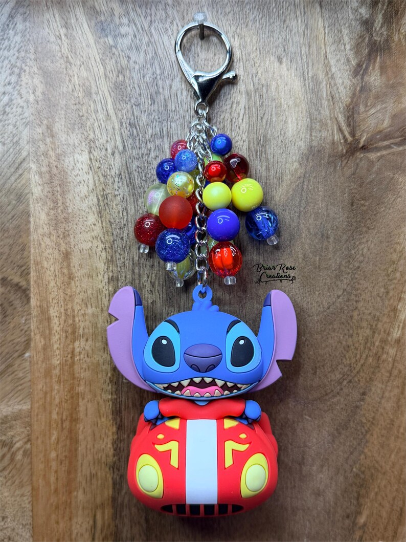 Puede incluir: Un llavero de Stitch azul con un coche rojo, sujeto a una cadena y cierre plateados. El llavero tiene un grupo de cuentas de colores rojo, amarillo, azul y transparente. El texto "Briar Rose Creations" es visible.