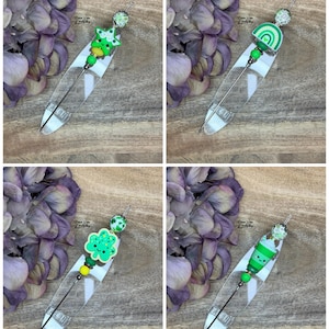 Peut inclure: Quatre marque-pages décoratifs en forme de plumes sur le thème de la Saint-Patrick. Chaque marque-page est orné d'une plume transparente, d'une épingle en métal et d'une perle décorative. Les motifs comprennent une étoile verte, un arc-en-ciel, un trèfle à quatre feuilles et un motif à rayures vertes.