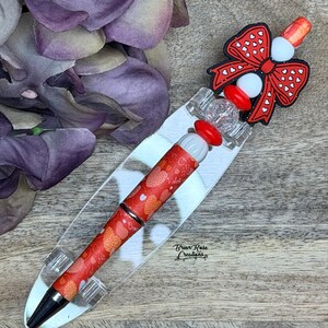 以下が含まれることがあります： ハート模様の赤いペン。透明なアクリルスタンドに展示されています。ペンは赤いリボンと白いビーズで飾られています。ペン先は黒で、バレンタインデーをテーマにしたデザインです。