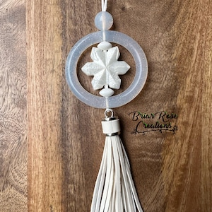 May include: White snowflake charm with a white tassel hanging from a clear, glittery circle. The charm is attached to a white cord.