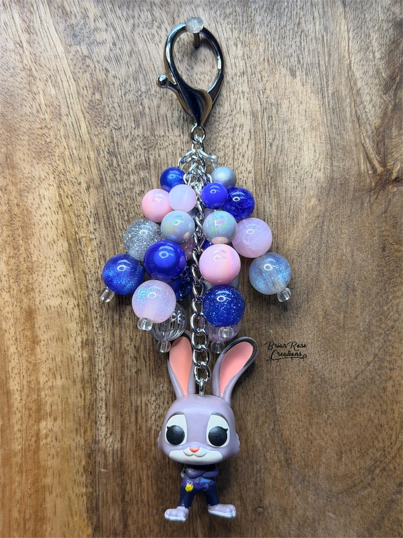 May include: A keychain featuring a purple bunny figurine with pink ears and a blue uniform. The keychain has a cluster of blue, pink, and silver beads, attached to a silver clasp. The text "Briar Rose Creations" is visible.