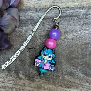 May include: A metal bookmark with a floral design and a charm. The charm features a blue and teal dragon reading a book, with purple and pink beads. The text "Briar Rose Creations" is visible.