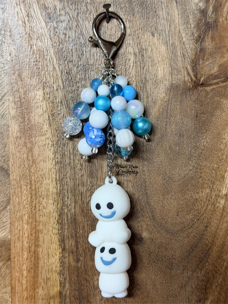 May include: A keychain featuring a silver-toned lobster clasp attached to a cluster of blue and white beads in various textures and finishes. Below the beads, two stacked white silicone snowman-like figures with blue smiles are suspended.