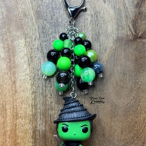 May include: A keychain with a green witch figurine in a black hat and dress, holding a broom. The charm is decorated with a cluster of green, black, and iridescent beads, connected to a silver-tone clasp.