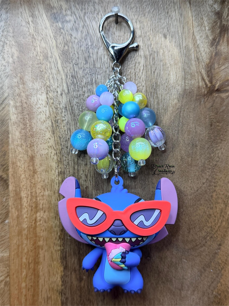 Puede incluir: Un llavero de Stitch azul con gafas de sol rojas y una bebida, sujeto a un cierre plateado y un grupo de cuentas de colores. El llavero est&aacute; sobre un fondo de madera.