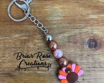 Op de afbeelding: Een bruine, oranje en roze kalkoenhanger met een zilveren sleutelhanger. De hanger is gemaakt van siliconen en heeft een glitterafwerking. De sleutelhanger is bevestigd aan een zilveren ketting met zilveren kralen.