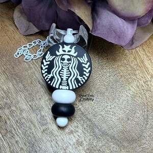 May include: A black and white badge reel with a skeleton design inside a circular logo. The badge reel has a white chain and a decorative beaded accent in white and black. The background includes purple flower petals and a wooden surface.