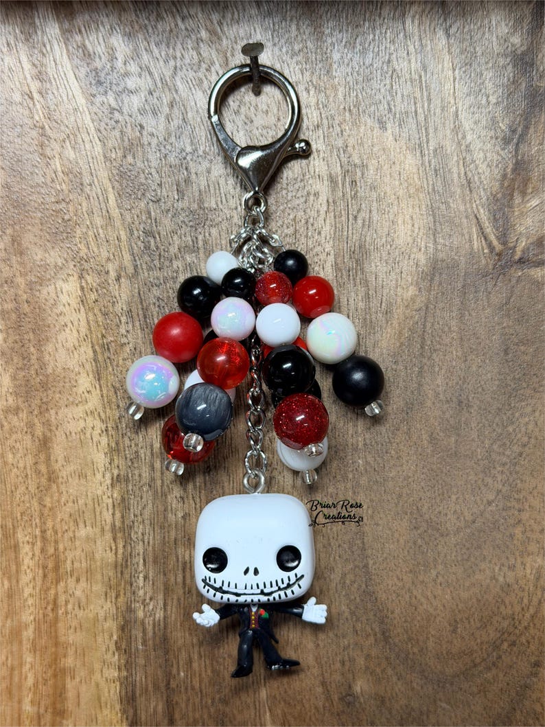 May include: A keychain featuring a silver-toned metal lobster clasp and chain adorned with a cluster of round beads in black, white, red, and iridescent finishes. A small vinyl figure of a skeleton character in a suit hangs at the bottom.