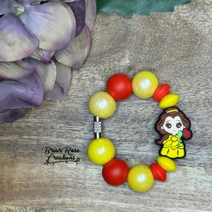 May include: A yellow and red beaded bracelet with a Belle charm. The charm features Belle from Beauty and the Beast holding a red rose.
