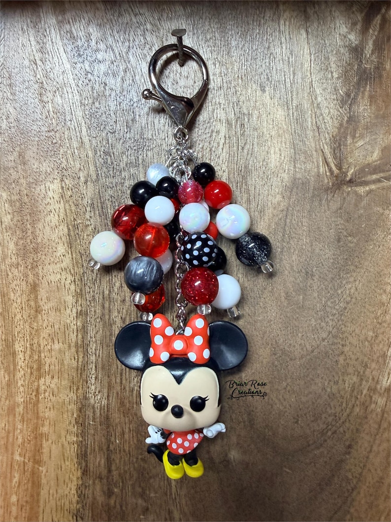 May include: A keychain featuring a stylized cartoon mouse figure with large black ears, a red and white polka dot bow, and a matching dress. The figure hangs from a silver metal clasp attached to a cluster of round beads in red, black, and white.