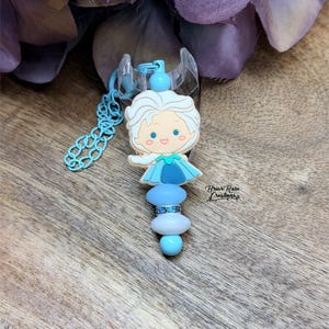 Puede incluir: Un clip para chupete azul y blanco con el tema de Elsa. El clip presenta una figura de dibujos animados de Elsa, cuentas azules y una cadena. El clip está unido a una pieza de plástico transparente. El texto "Briar Rose Creations" es visible.