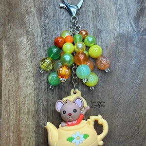 Pode incluir: Um porta-chaves com um berloque de bule amarelo e um personagem de rato. O berloque é adornado com um grupo de contas coloridas em tons de verde, laranja e amarelo. O porta-chaves tem um fecho prateado.