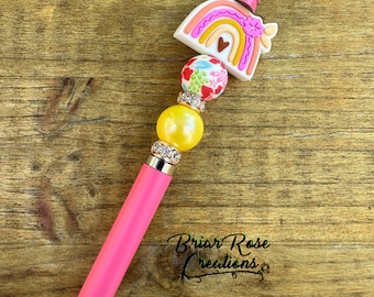 Puede incluir: Bolígrafo de estilo rosa con un diseño de arcoíris y flores. El bolígrafo tiene un cuerpo rosa y una parte superior de arcoíris con un corazón en el centro. La parte superior está decorada con un diseño floral y una cuenta rosa. El bolígrafo está rematado con una cuenta transparente y una cuenta dorada.