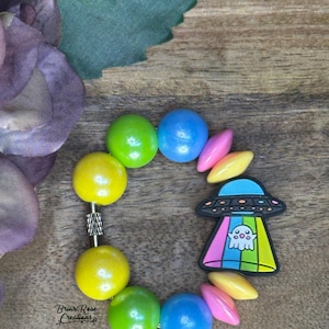 Puede incluir: Un brazalete de cuentas de colores con un amuleto de OVNI de colores arcoíris que presenta un fantasma en su interior. El brazalete está hecho con cuentas de madera redondas en tonos verdes, amarillos, azules y rosas.