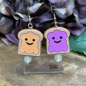 May include: A pair of earrings featuring a purple and orange peanut butter and jelly sandwich design. The earrings are made of silicone and have a shiny, white bead on the bottom of each earring.