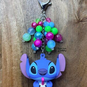 Puede incluir: Un llavero de Stitch azul con un grupo de cuentas coloridas. El llavero presenta un personaje de dibujos animados sosteniendo una bebida con una pajita rosa. Las cuentas son de varios colores, incluyendo rosa, azul y verde, y est&aacute;n unidas a un cierre plateado.