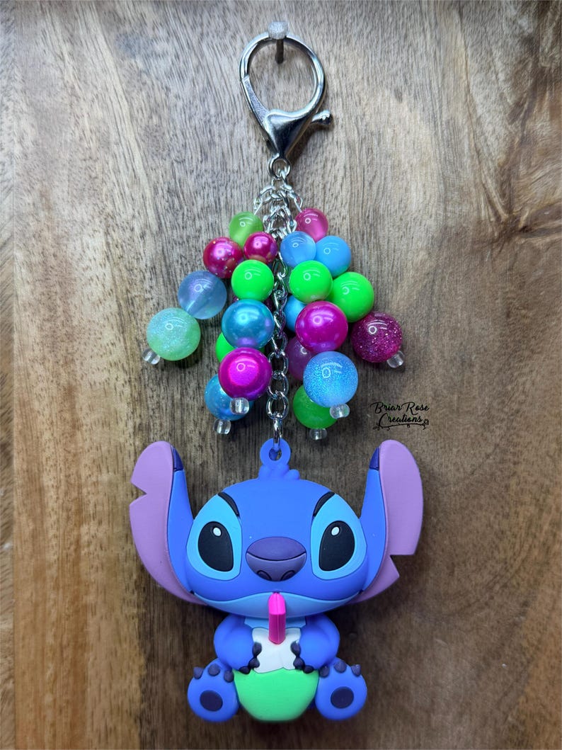 Puede incluir: Un llavero de Stitch azul con un grupo de cuentas coloridas. El llavero presenta un personaje de dibujos animados sosteniendo una bebida con una pajita rosa. Las cuentas son de varios colores, incluyendo rosa, azul y verde, y est&aacute;n unidas a un cierre plateado.