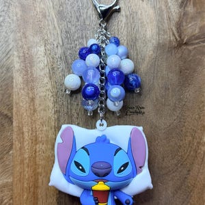 Puede incluir: Un llavero de Stitch azul con un grupo de cuentas azules y blancas. Stitch sostiene una taza roja y amarilla mientras descansa sobre una almohada blanca. El llavero tiene un cierre y una cadena plateados.