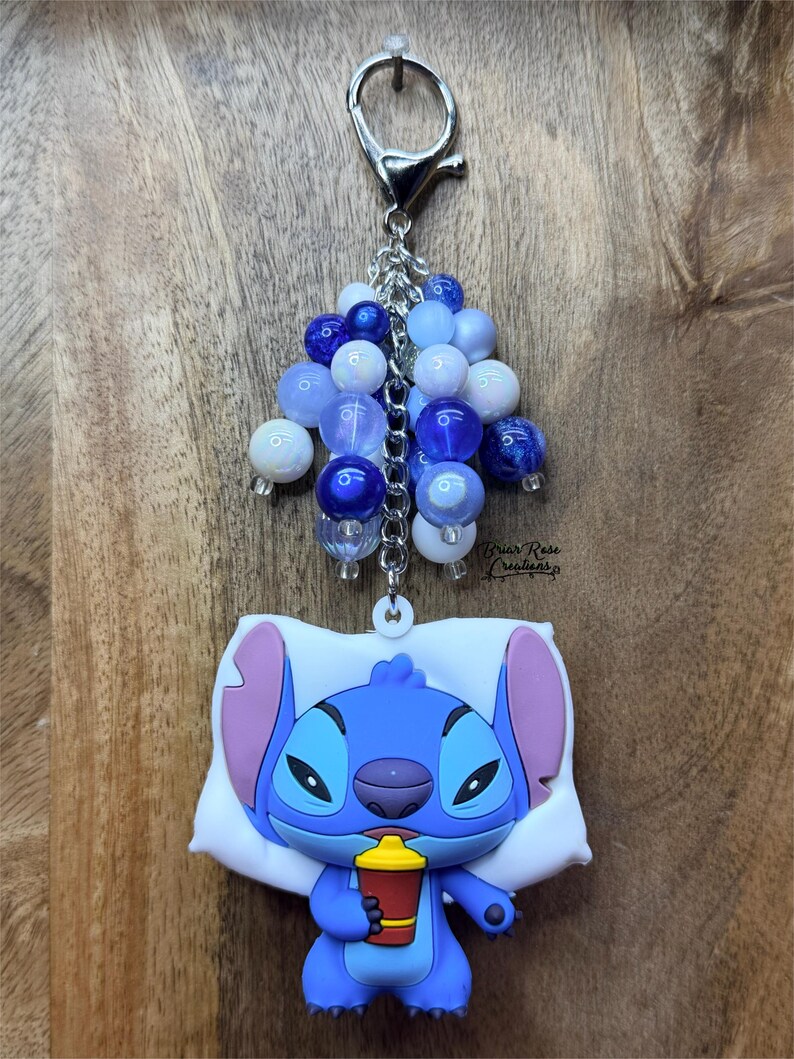 Puede incluir: Un llavero de Stitch azul con un grupo de cuentas azules y blancas. Stitch sostiene una taza roja y amarilla mientras descansa sobre una almohada blanca. El llavero tiene un cierre y una cadena plateados.