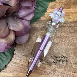 Puede incluir: Pluma morada y plateada con un encanto de murciélago brillante. La pluma está en un soporte de acrílico transparente. El texto "Briar Rose Creations" es visible en la esquina inferior derecha de la imagen.