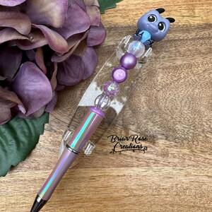 May include: A purple and iridescent pen with a blue and purple character on top. The pen is decorated with purple and clear beads. The pen is on a clear acrylic stand. The text "Briar Rose Creations" is visible in the image.