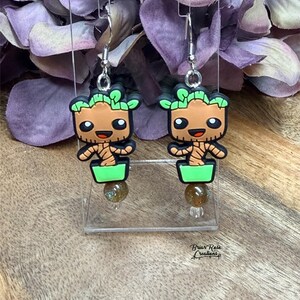 Könnte beinhalten: Ohrringe zum Aufhängen mit einem Cartoon-Charakter-Design. Jeder Ohrring hat ein braunes Gesicht, grüne Blätter und einen grünen Topf. Unter jedem Charakter befindet sich eine runde, gesprenkelte Perle. Die Ohrringe hängen an silberfarbenen Haken.
