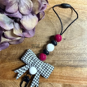 May include: A keychain accessory featuring a black and white houndstooth fabric bow, a white silicone bead, and a black metal lobster clasp. The cord includes a sequence of two magenta, two black, and one white silicone beads. Text reads Briar Rose Creations.