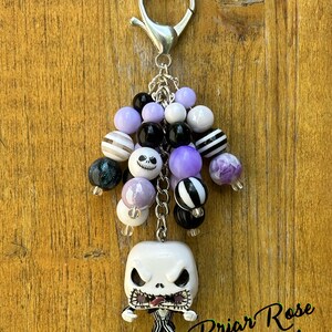 May include: A purple, black, and white beaded keychain with a Jack Skellington character charm. The keychain has a silver lobster clasp and a silver chain. The beads are a mix of solid colors, stripes, and patterns. The charm is a small, detailed figure of Jack Skellington from The Nightmare Before Christmas.