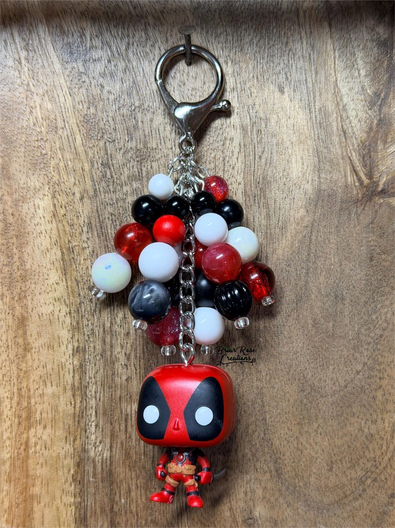 May include: A keychain featuring a small red and black character figurine with a large head and a cluster of round beads in red, white, black, and grey. The beads are attached to a silver-toned metal chain and a lobster claw clasp at the top.