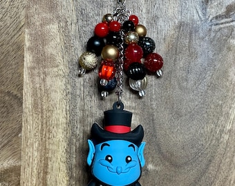 May include: A blue genie character keychain with a red cape, black top hat, and gold lamp. The keychain has a silver chain with red, black, and gold beads.